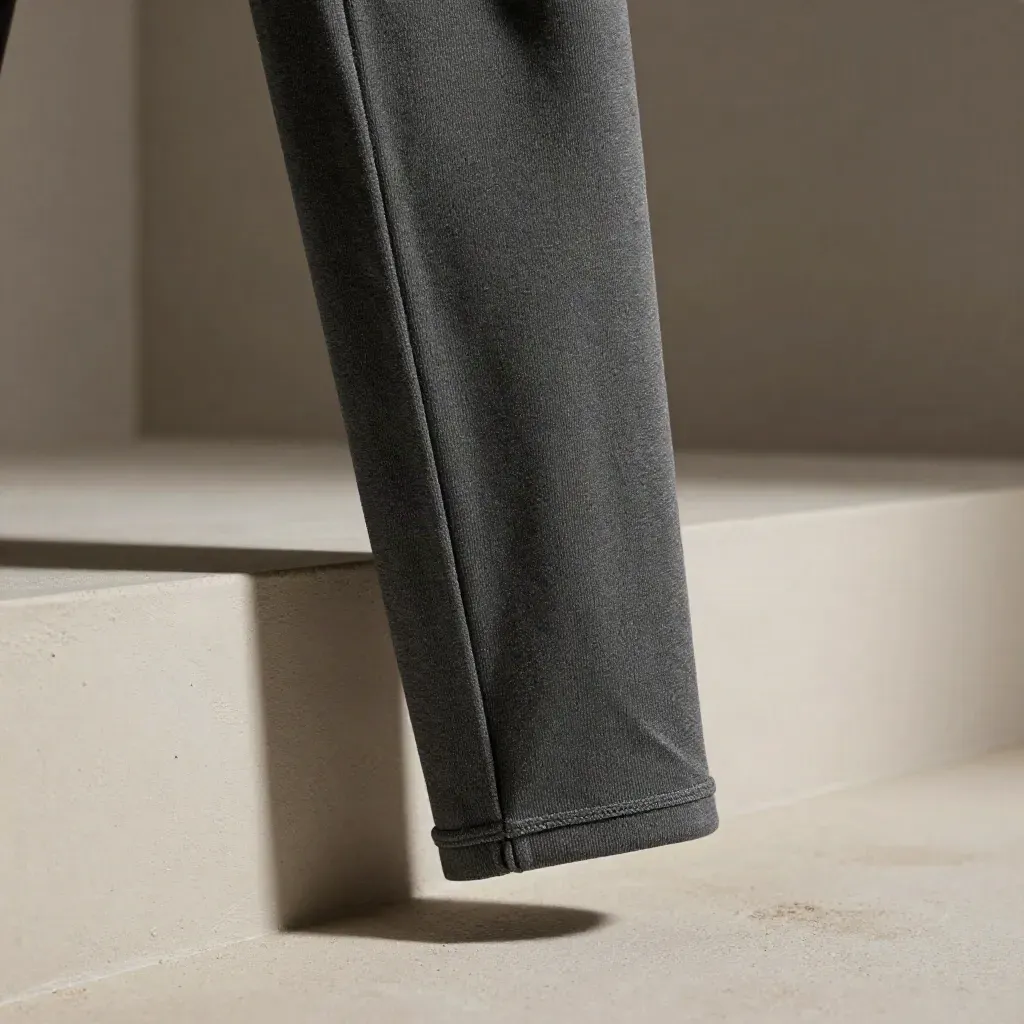 Charcoal jersey pant leg draped over a plaster rail, macro texture and hem detail in raking light.